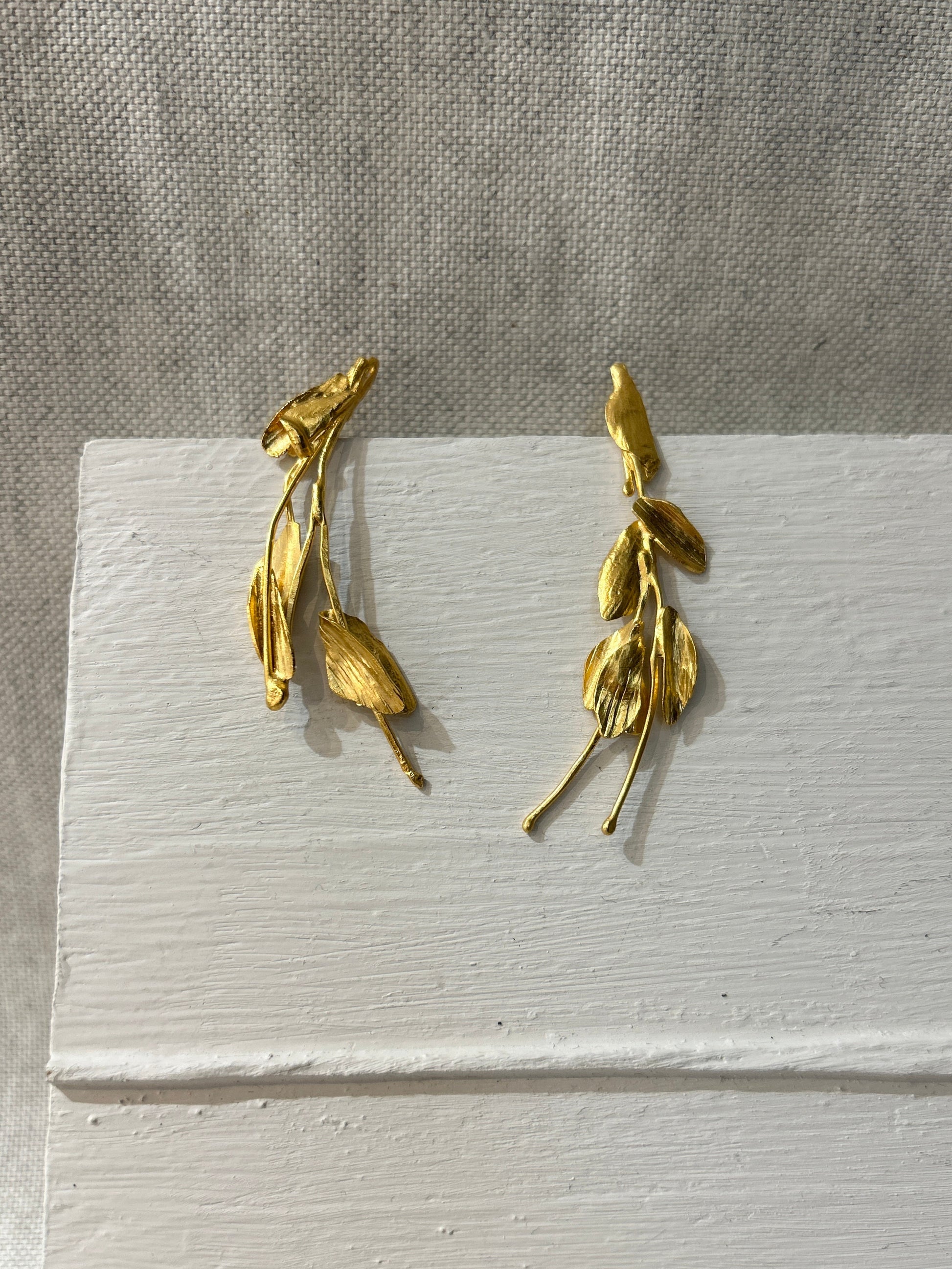 Gold leaf-shaped earrings on a white card with a gray background