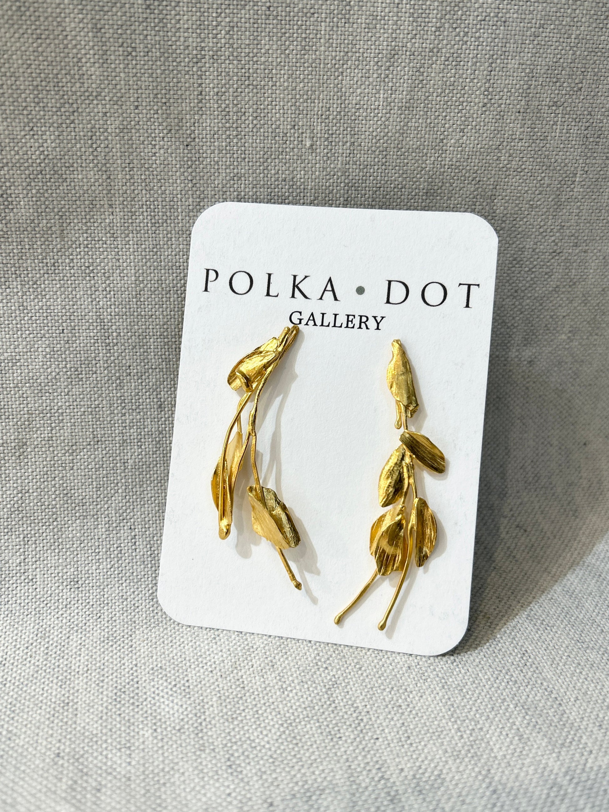 Gold leaf-shaped earrings on a Polka Dot Gallery card against a gray fabric background